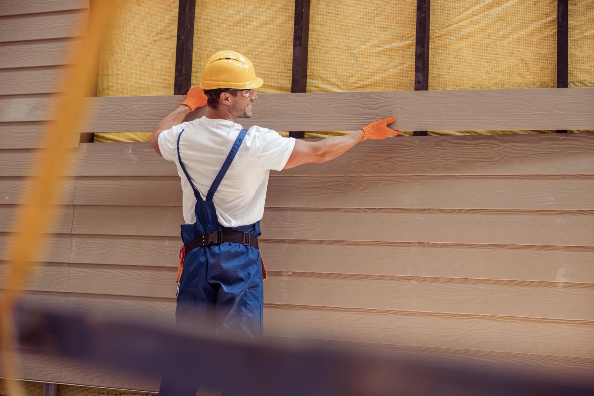 A smiling male worker is installing vinyl siding on a cabin under construction.