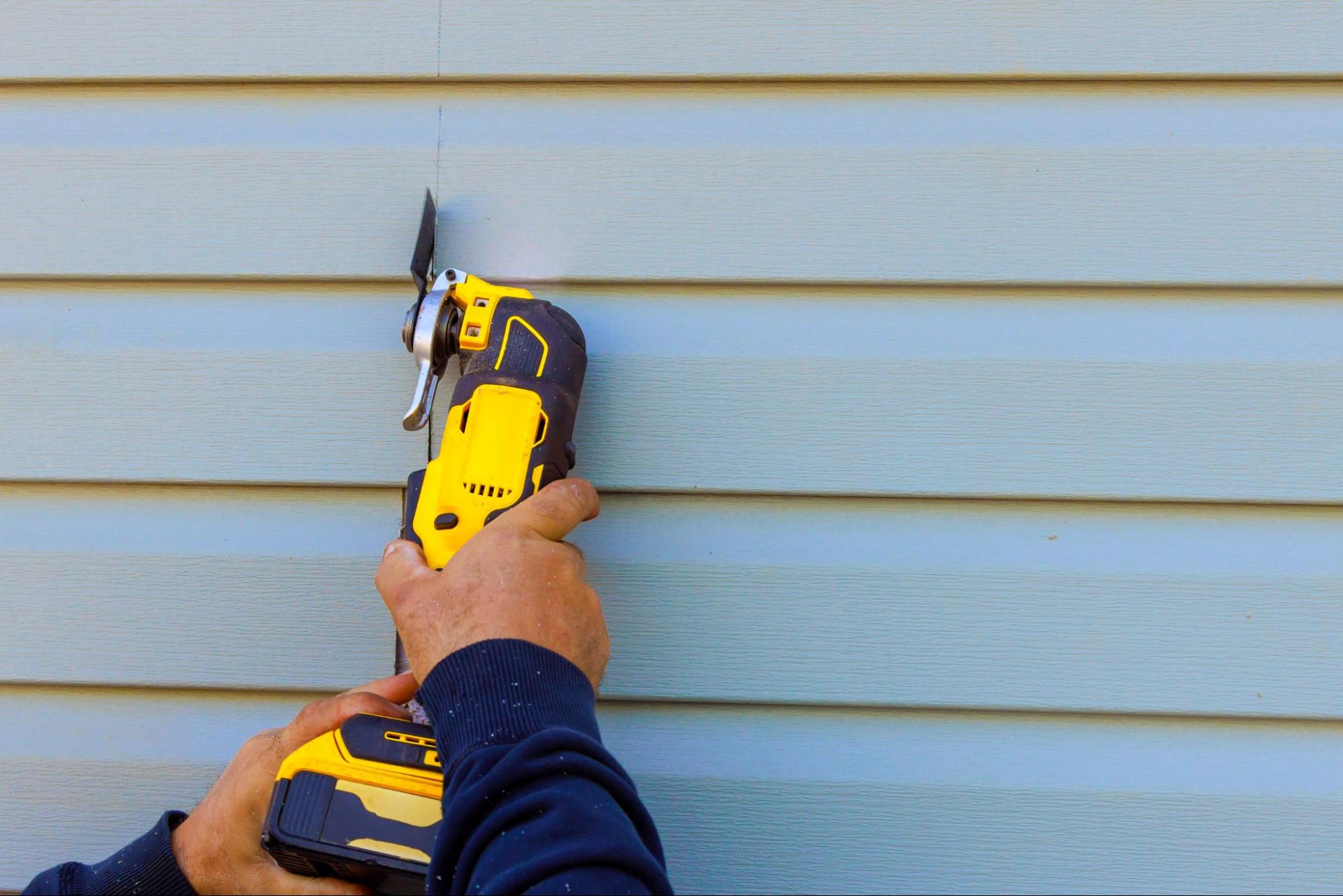 Worker removing cracked vinyl siding with a multi-tool during renovation.