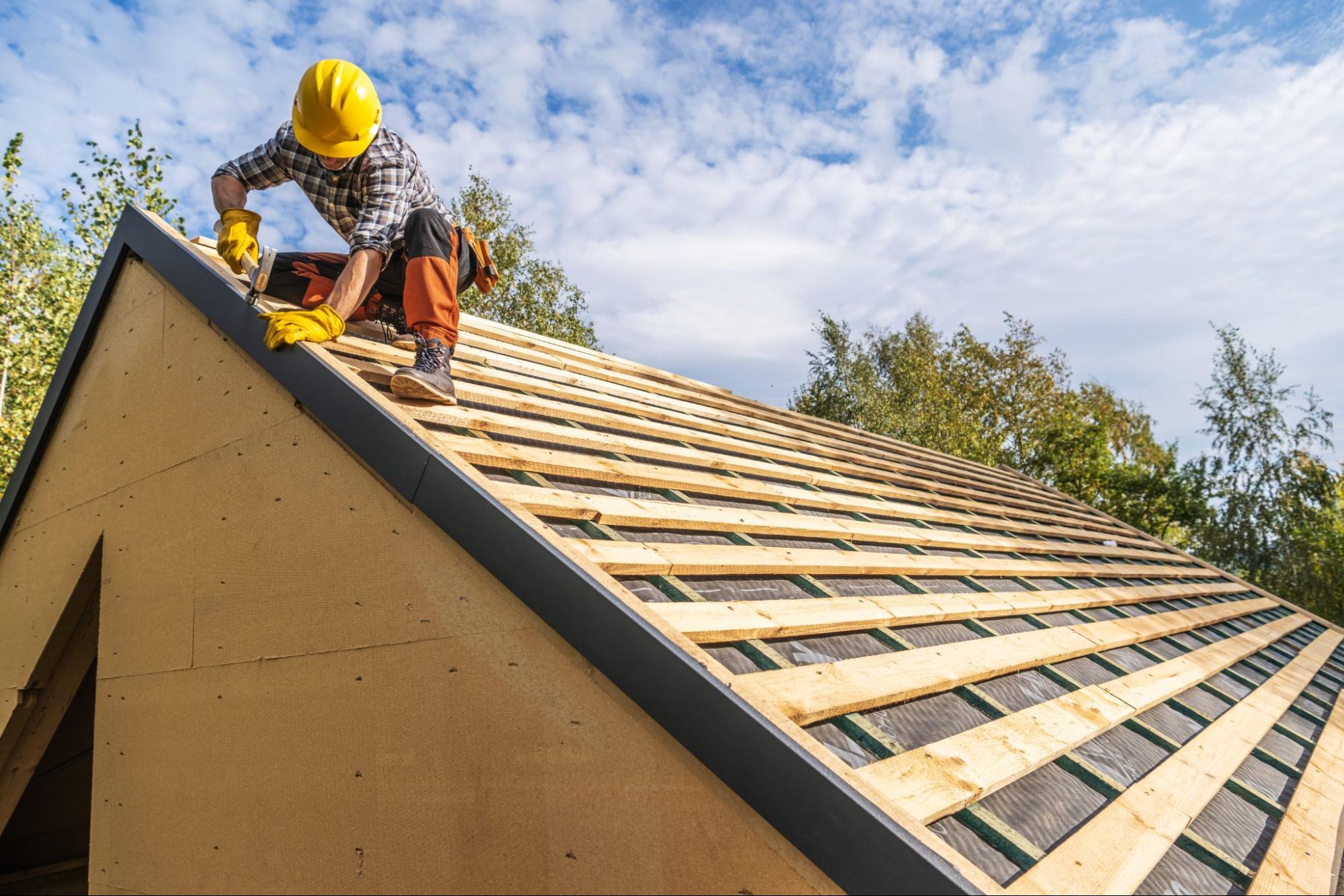 A roofer removes old asphalt shingles and carefully places the roofing materials into a blue container.