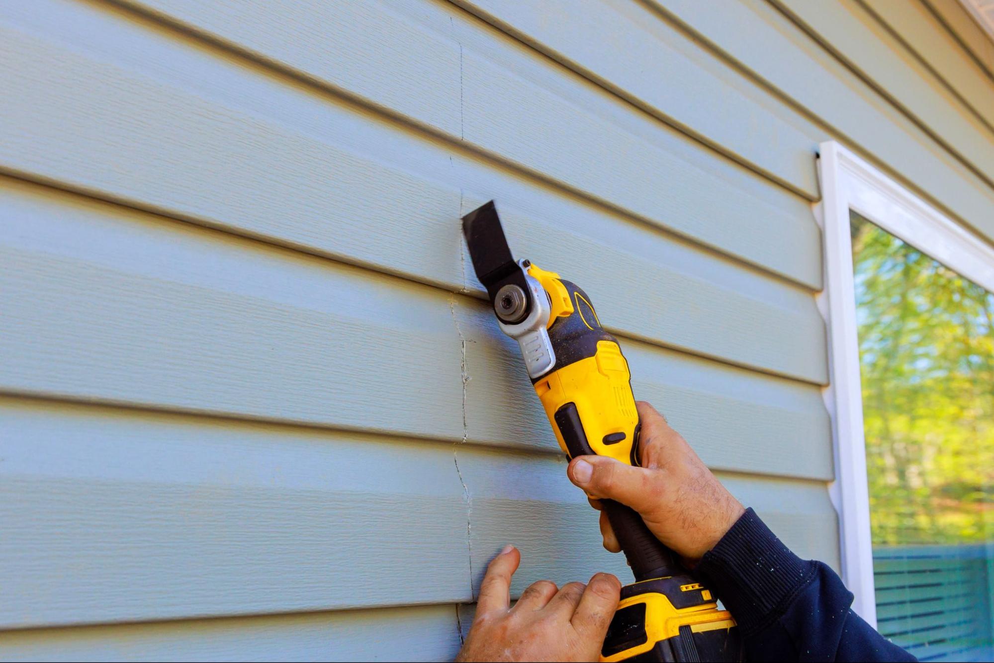 A close-up of a repairman's hands removing the old siding using a special tool to prepare for replacement.