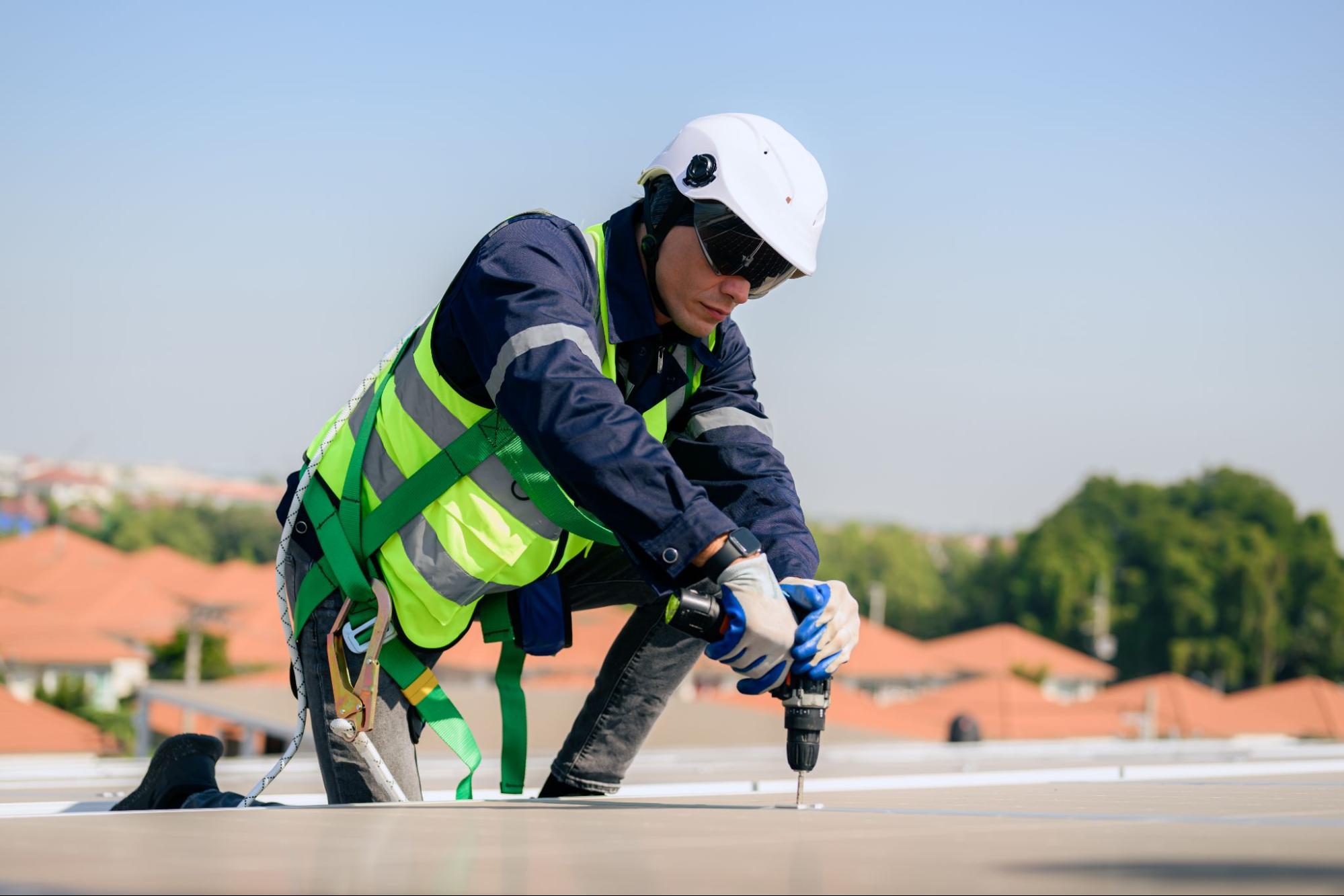 A roof repair specialist wearing a white hard hat and safety gear fixes a minor roof damage using a drill.