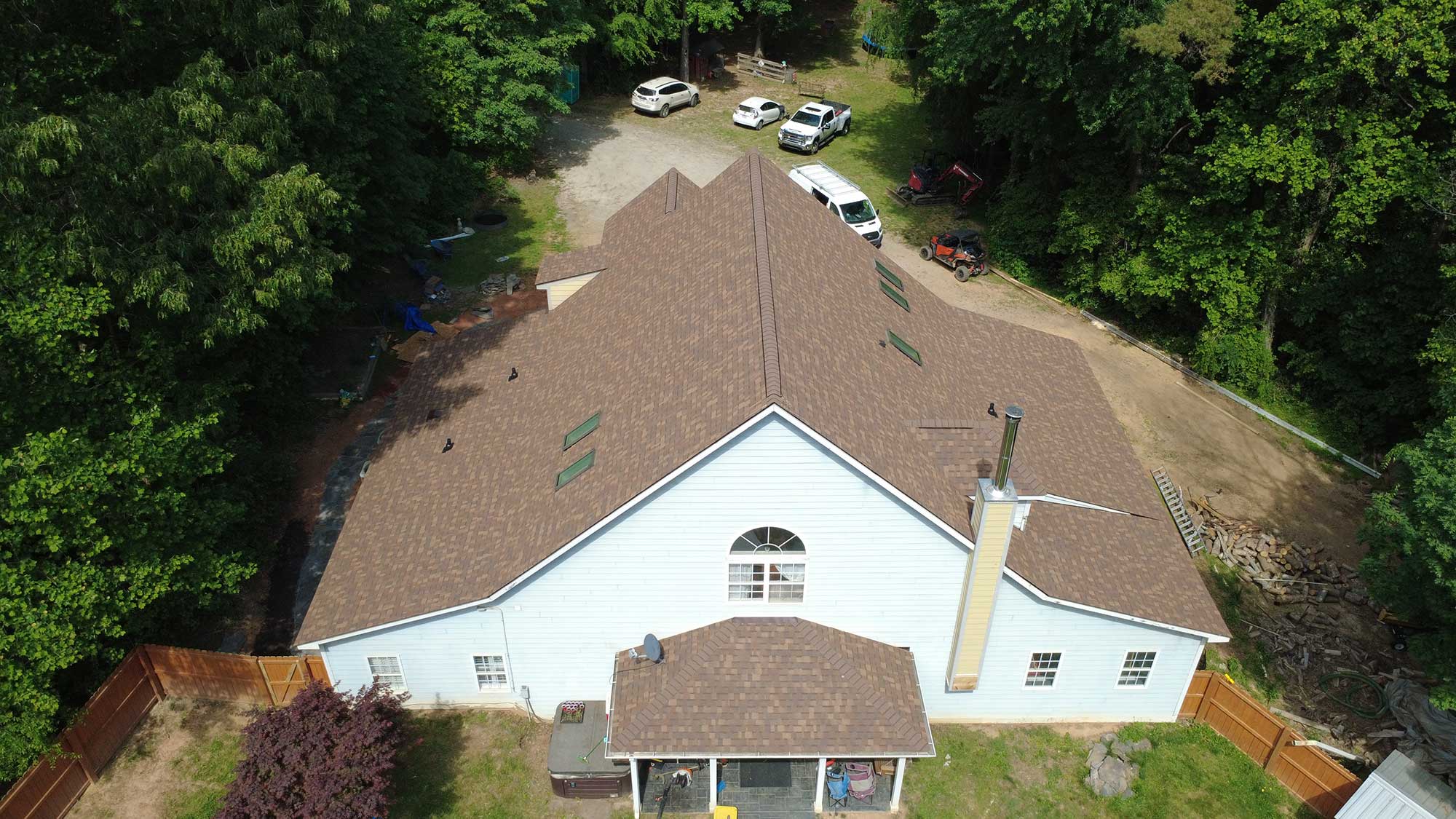 Aerial view of a large, light blue house with a newly installed brown shingle roof, surrounded by trees, a fenced yard, and several parked vehicles.