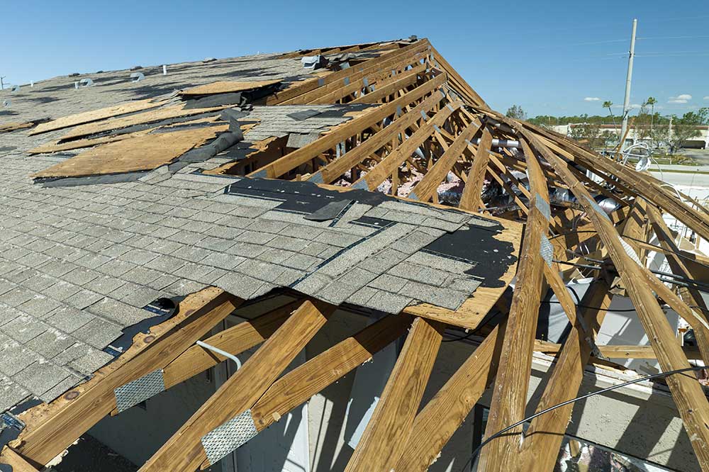 Damaged roof with missing shingles and exposed wooden beams against a clear sky.