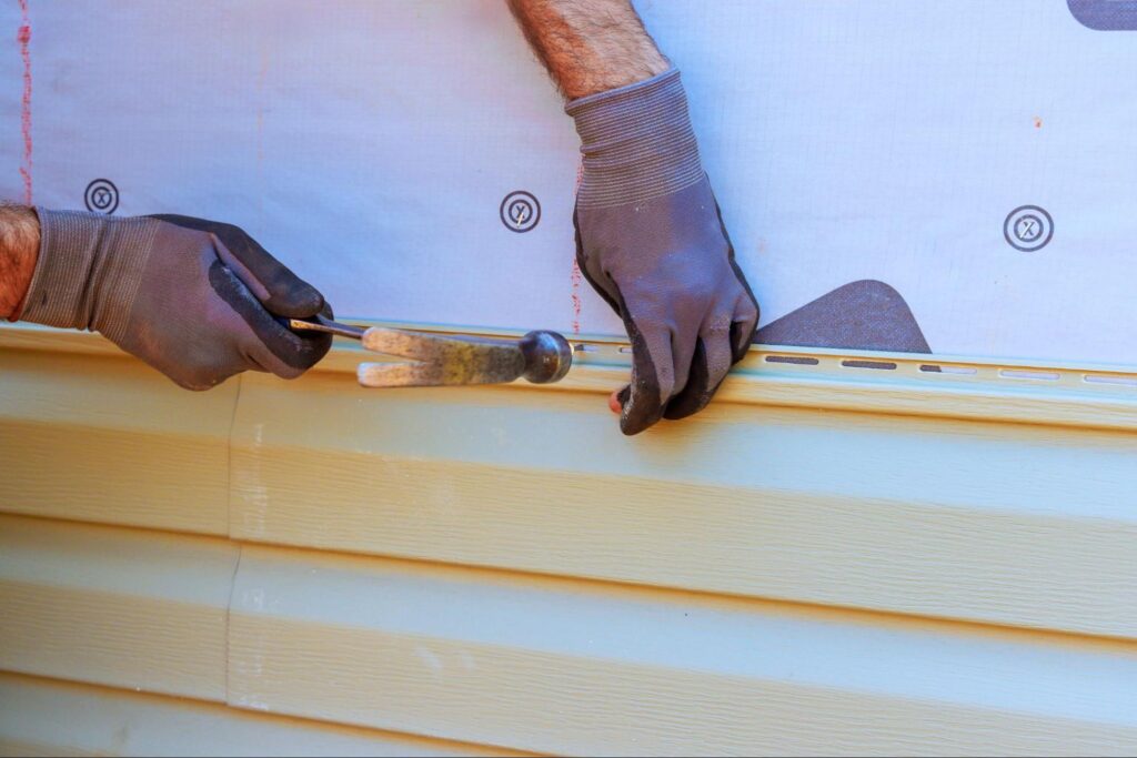 Close-up of a contractor’s hands installing vinyl siding with a hammer and level.