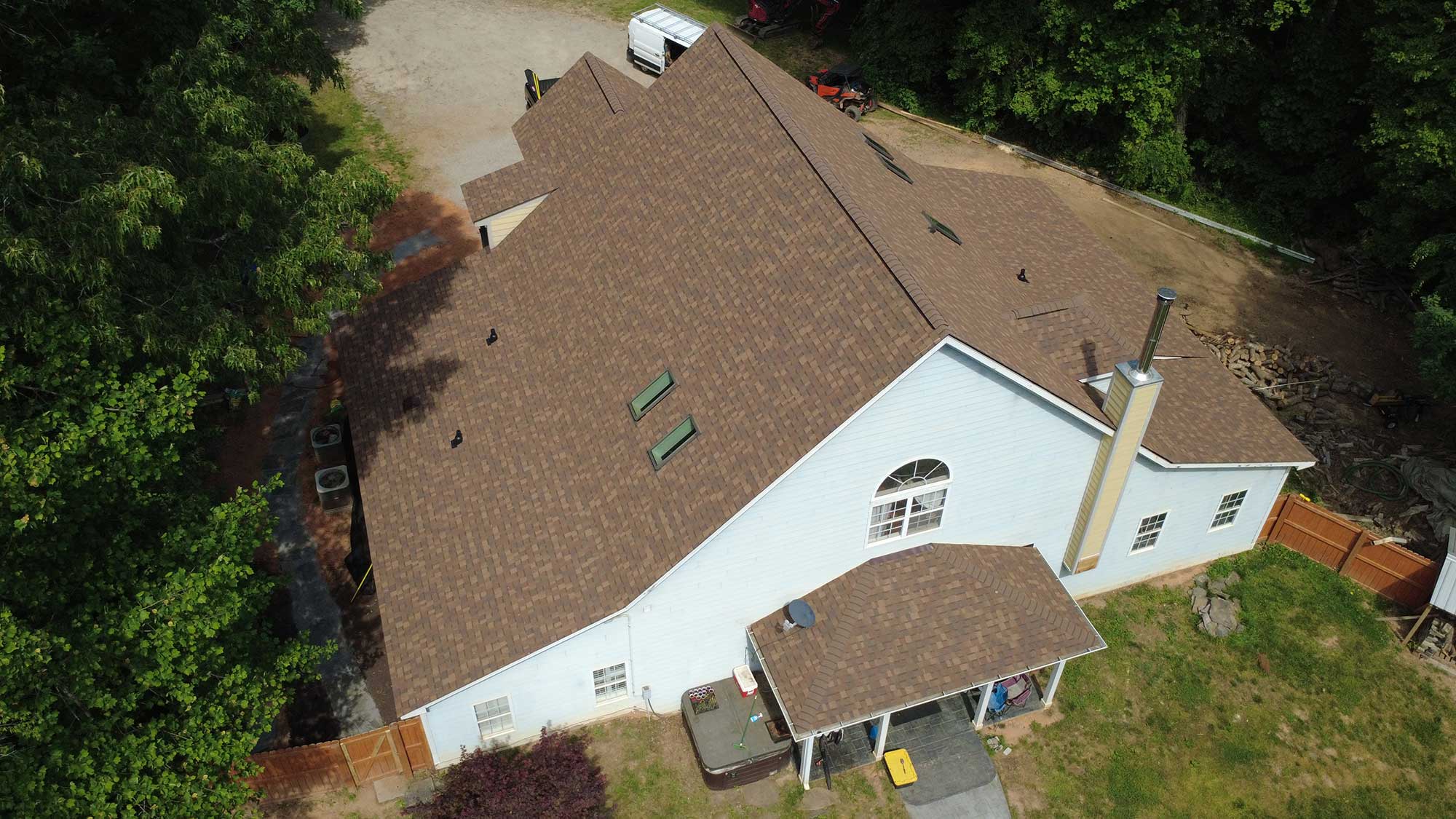 light blue house with a newly installed brown shingle roof, surrounded by trees, a fenced yard,