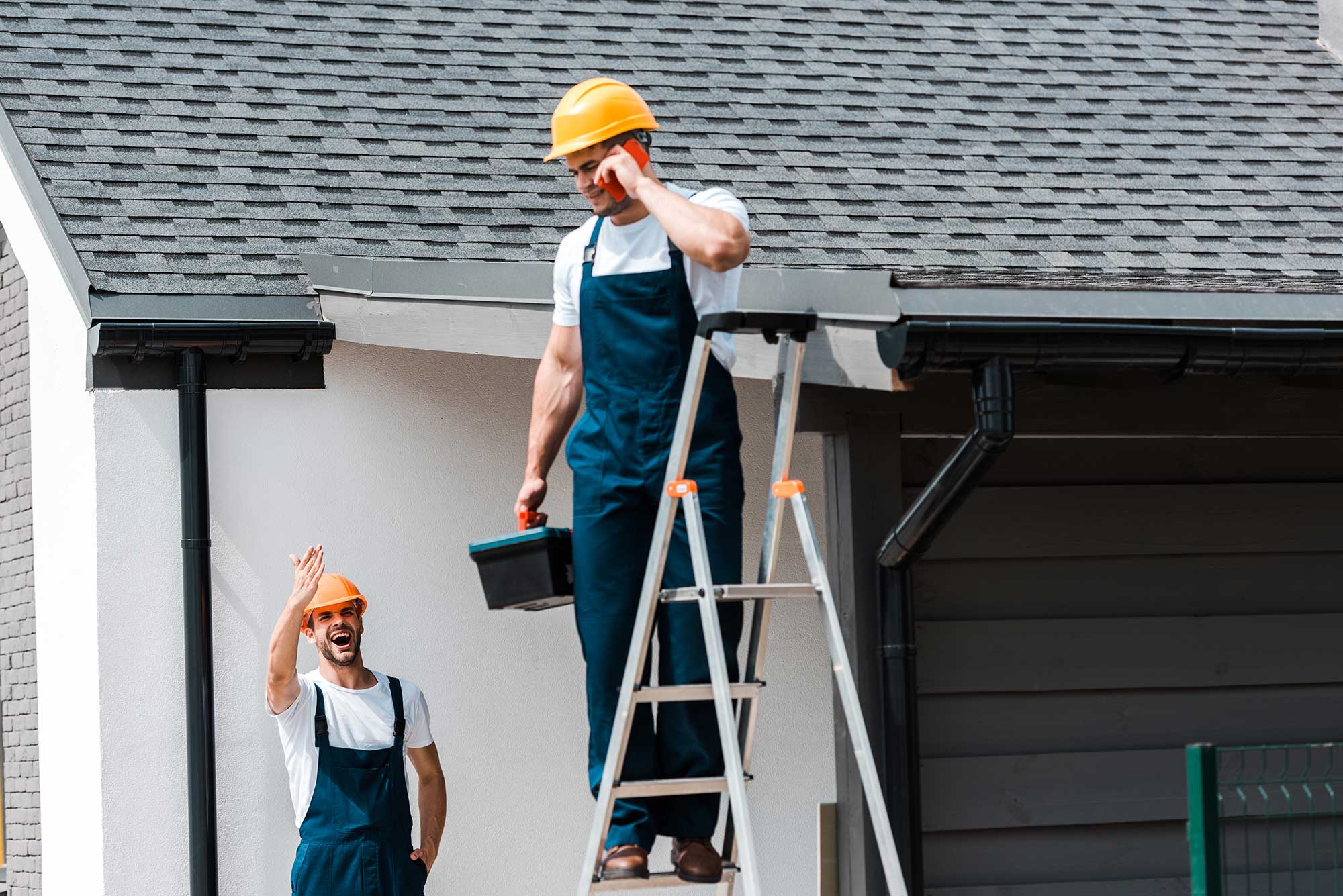 Two workers in overalls and hard hats, one on a ladder and another gesturing with hand.